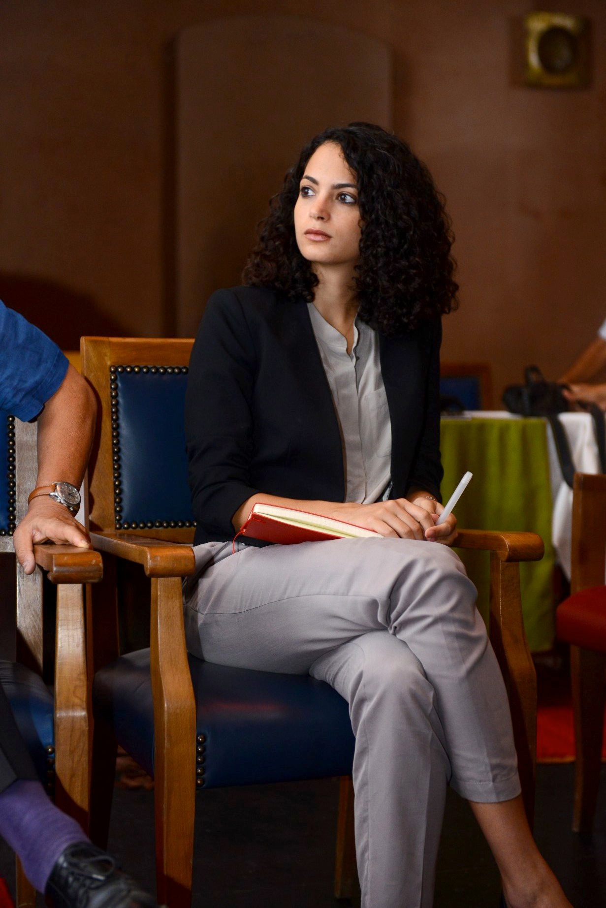Woman with curly dark hair sitting in wooden chair holding a red book, wearing black blazer and light pants.