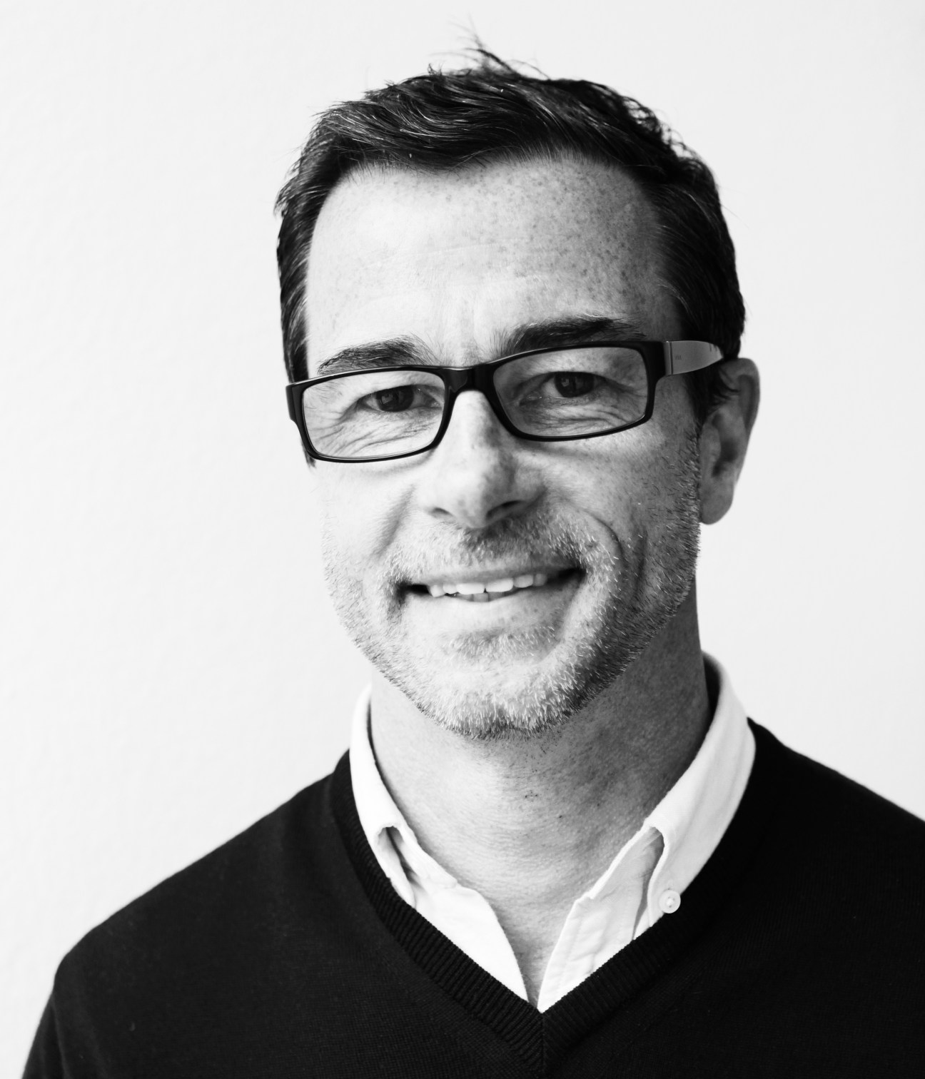 Black and white portrait photograph of a man wearing glasses and dark sweater over white shirt