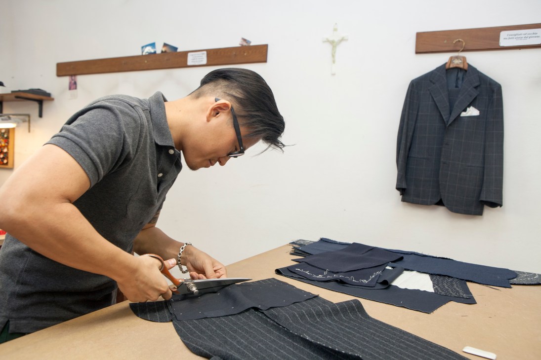 Hojun Choi, a tailor apprentice, carefully cuts dark fabric with scissors at a wooden workbench in the Liverano & Liverano atelier.