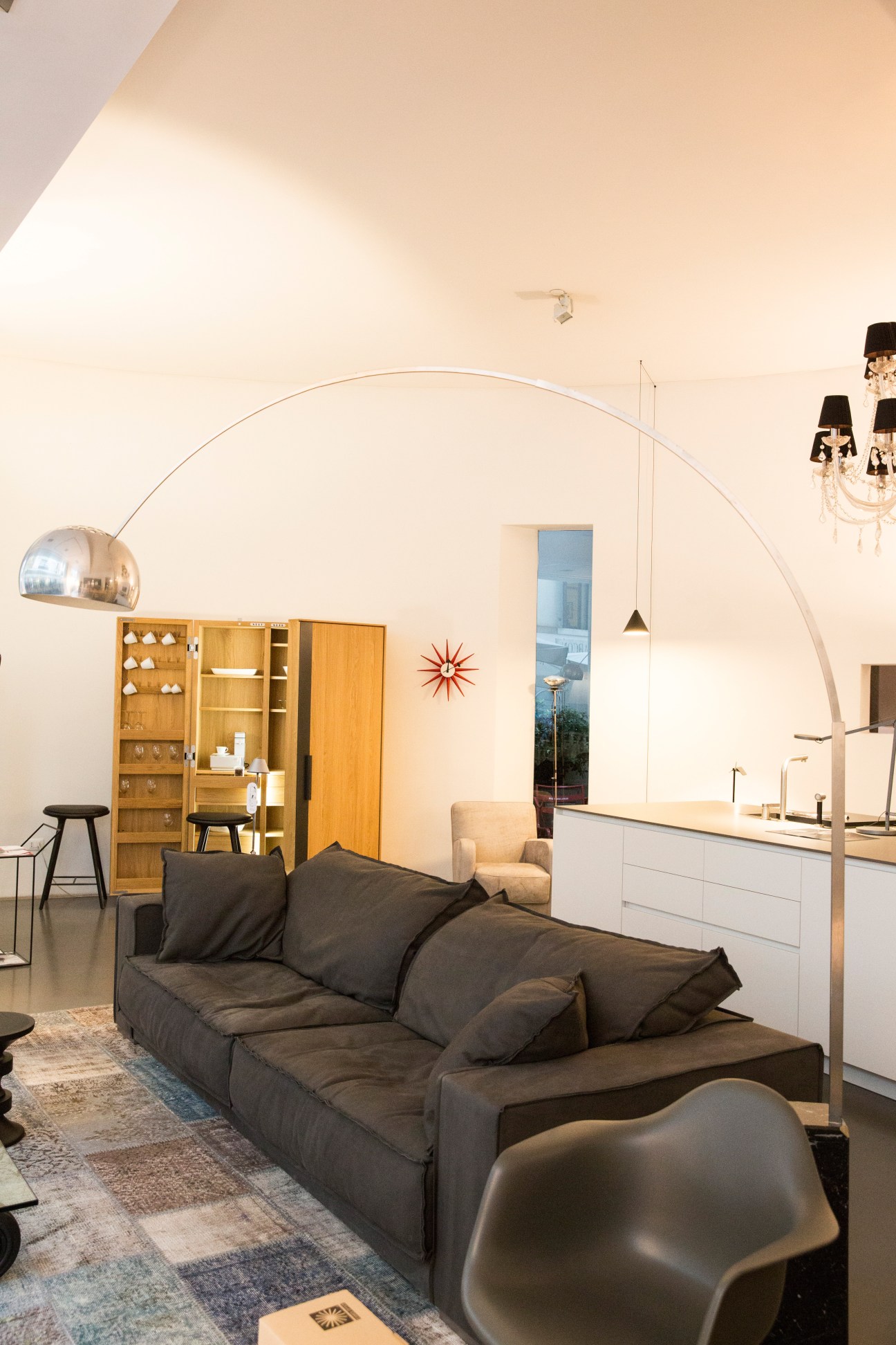 Modern furniture showroom displaying a dark sofa, arc lamp, wooden storage units, and design pieces in a minimalist space.