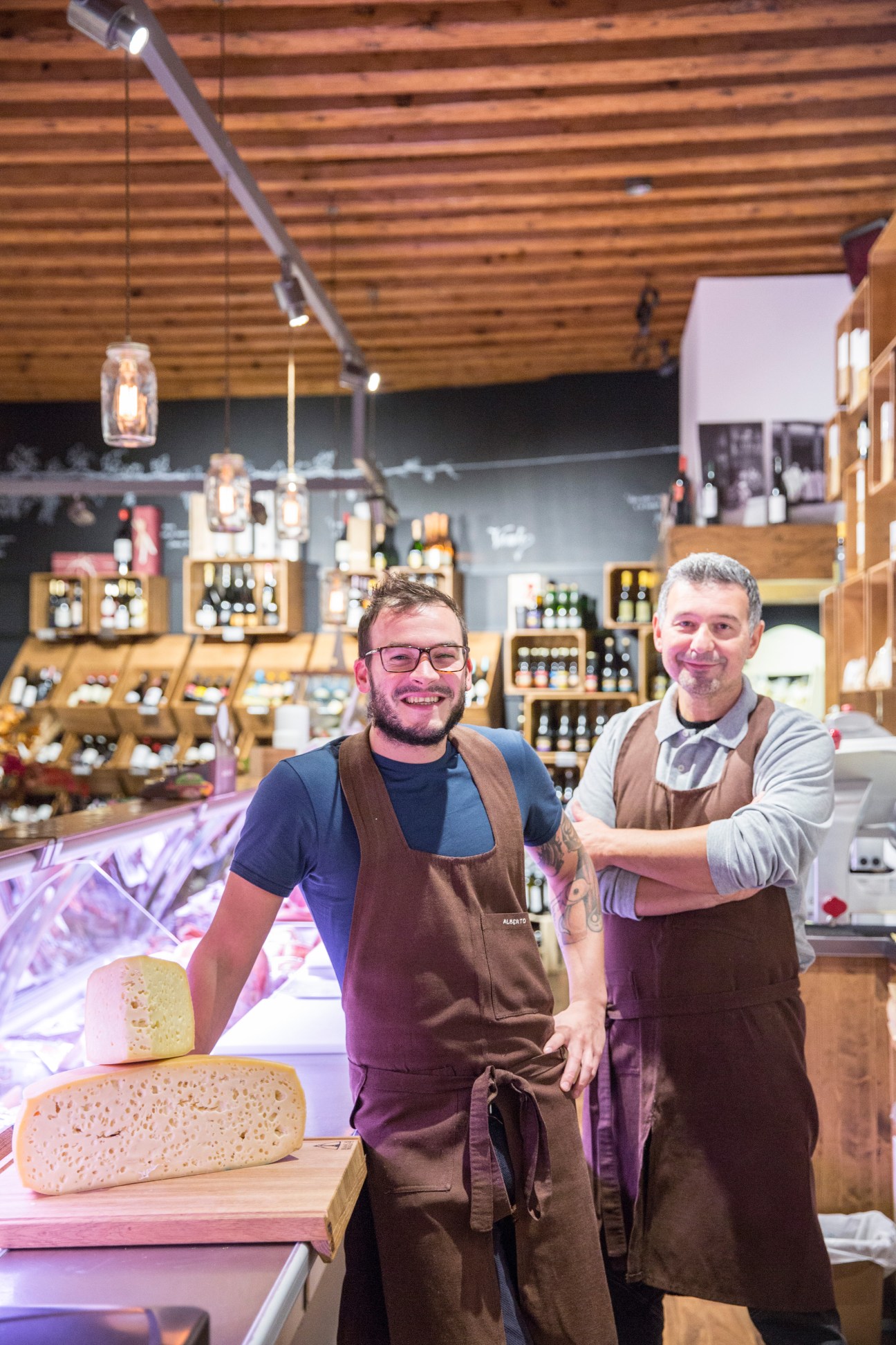 Two men in brown aprons stand behind a deli counter with sliced cheese and bottles of wine and beer displayed behind them.