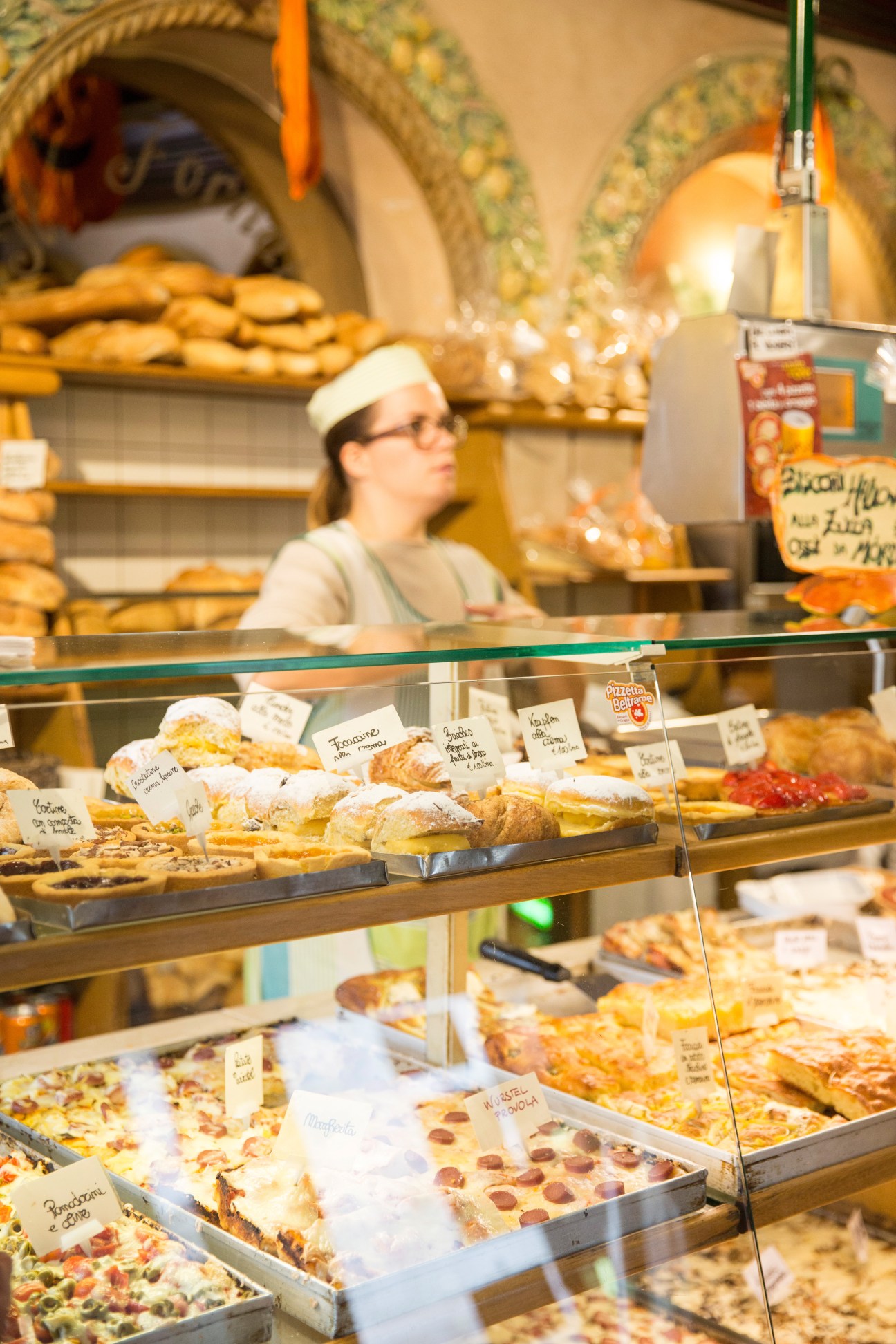 Bakery counter display with pastries, breads and baked goods behind glass, staff member in white hat visible in background.