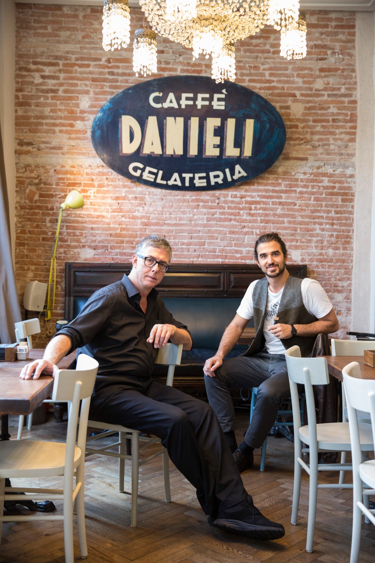 Two men sit in Danieli restaurant under a vintage oval sign reading 'Caffé Danieli Gelateria' with exposed brick walls and crystal chandeliers above.