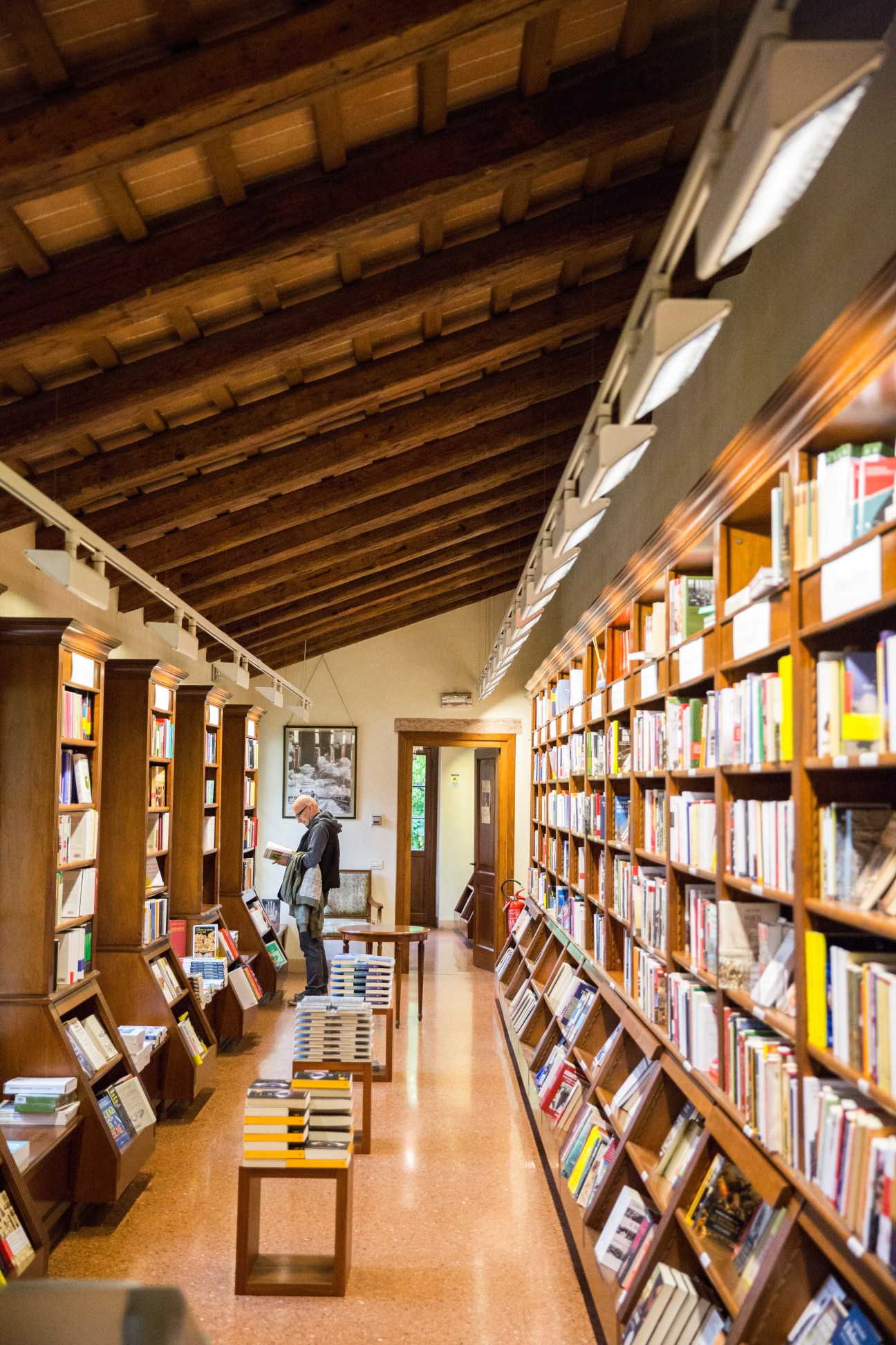 Interior of Libreria Palazzo Roberti bookshop with wooden beamed ceiling, walnut shelves, and books displayed on chairs.