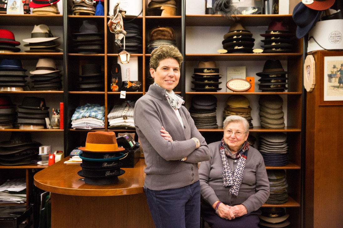 Two women stand in front of wooden shelves displaying stacked Borsalino hats and other hat styles in a boutique shop.