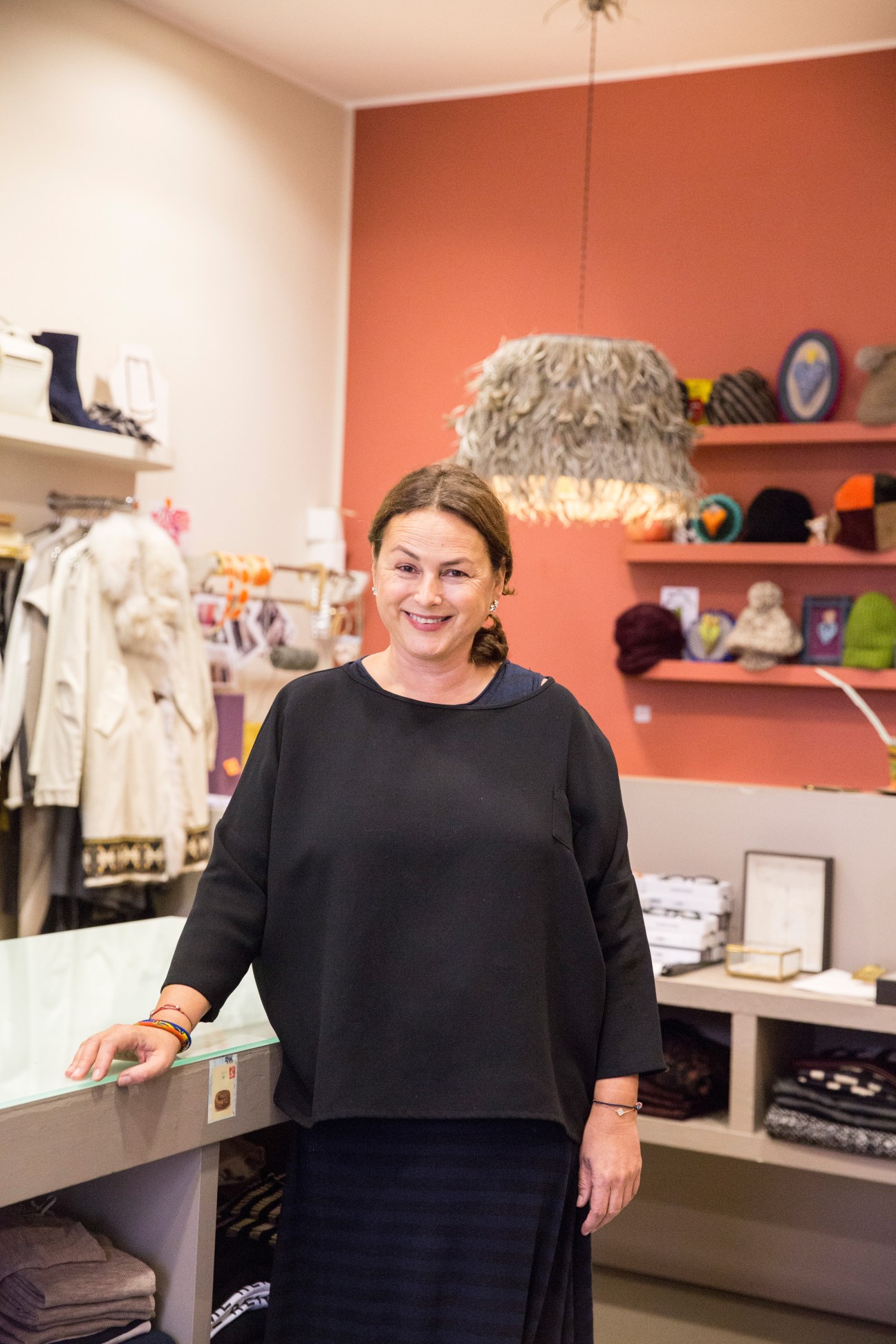 Woman standing in Zoe boutique with orange wall, clothing racks and shelves of merchandise behind her