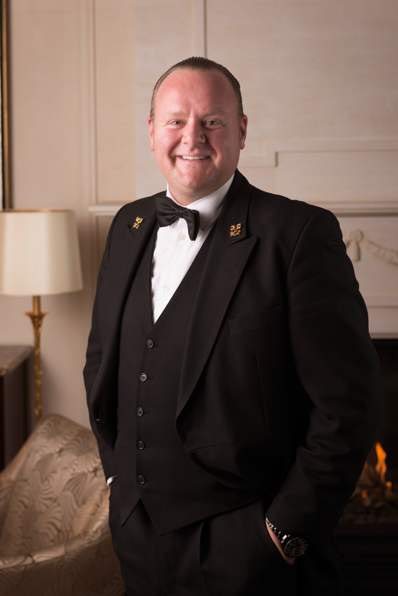 Man in formal black tuxedo with bow tie and gold buttons smiling in an elegant interior setting.