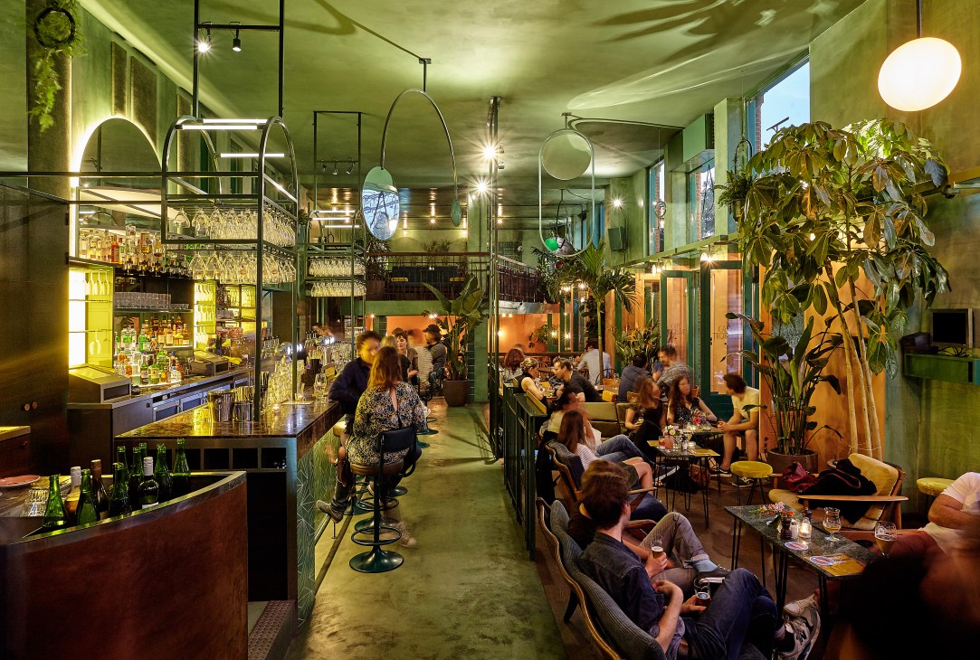 Bar Botanique interior with lush plants, art deco green tiles, suspended mirrors, and patrons dining among ferns and palm trees.