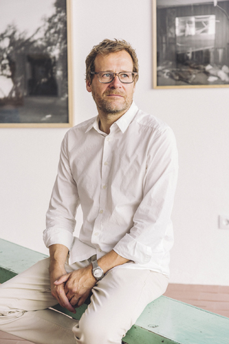 Man in white shirt and glasses sitting on bench with framed photographs on wall behind him.