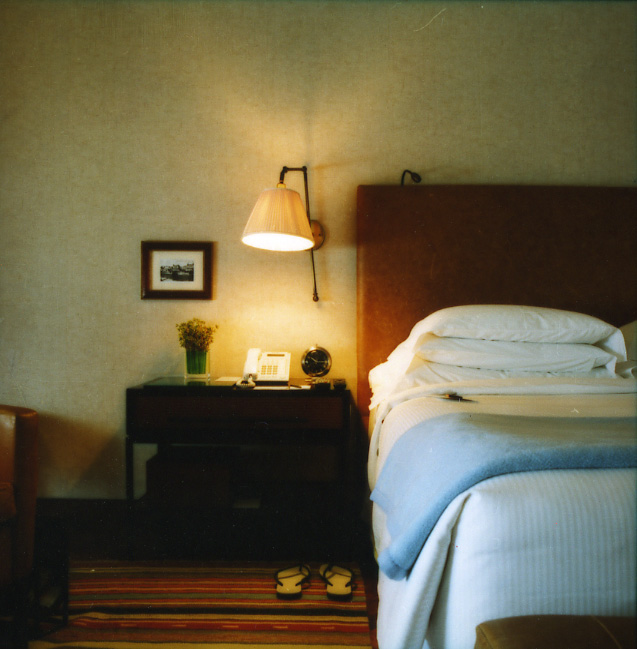 Hotel bedroom with wooden headboard, white bedding, dark nightstand, warm pendant lamp, and yellow slippers on wood flooring.