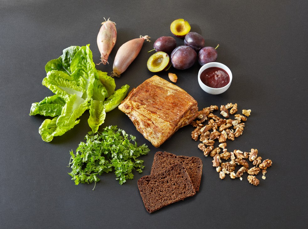 Rye bread slices with ingredients for pork belly smørrebrød including lettuce, shallots, plums, walnuts and plum purée on dark background.