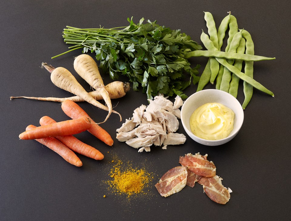 Ingredients for chicken salad with baked root vegetables including carrots, parsnips, snap peas, fresh herbs, shredded chicken, mayonnaise, bacon and curry powder arranged on a dark surface.