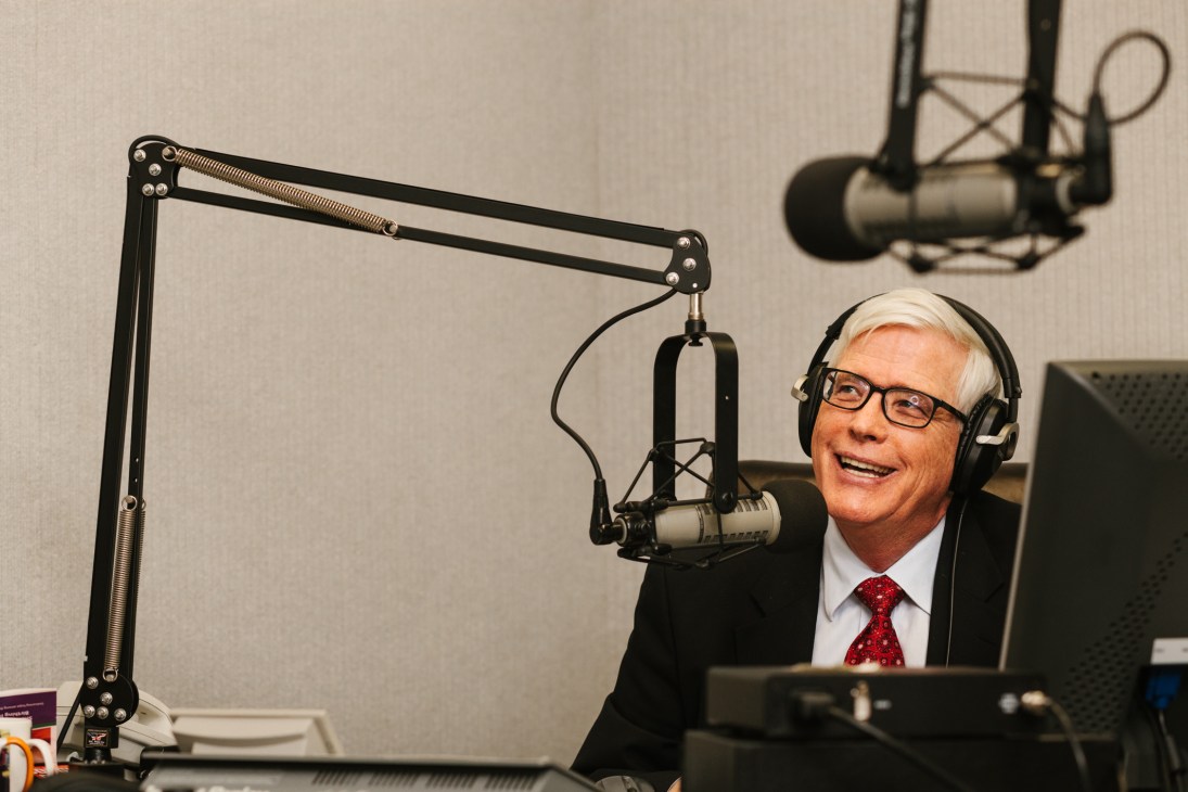 Hugh Hewitt, conservative radio host, wearing glasses and headphones at a microphone in a professional broadcast studio.