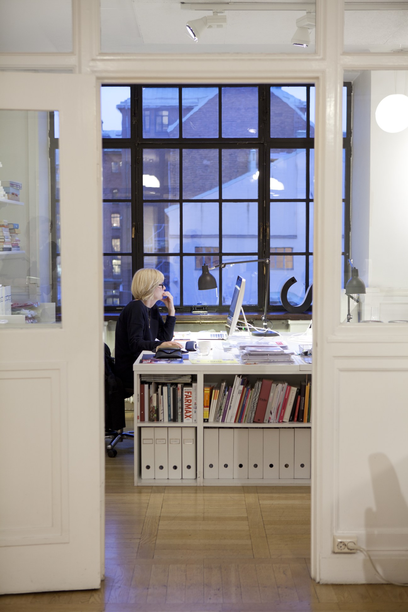 Woman working at desk in minimalist Stockholm office with black-framed windows and storage shelving.