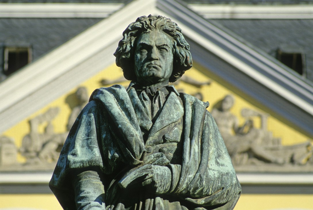 Bronze statue of Beethoven with long hair and period clothing, holding a scroll, with a yellow building facade in background.