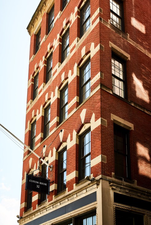 Red brick building facade with cream stone detailing and black-framed windows, with a sign reading 'Custom House 32'.
