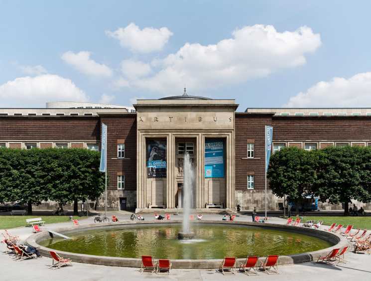 Neoclassical museum building with brick wings, central portico with fountain and red deck chairs in foreground.