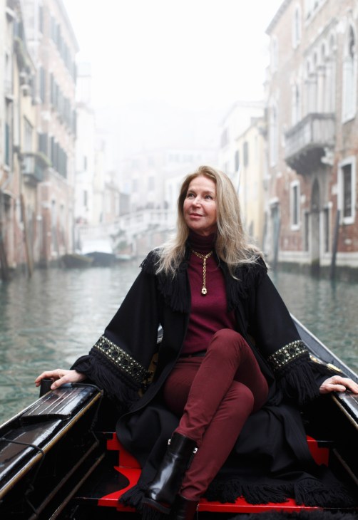 Woman in burgundy outfit and black coat sitting in a gondola on a Venice canal.