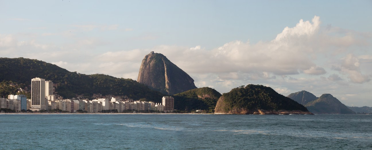 Rio de Janeiro's coastal cityscape with Sugarloaf Mountain and surrounding peaks visible across the blue water.