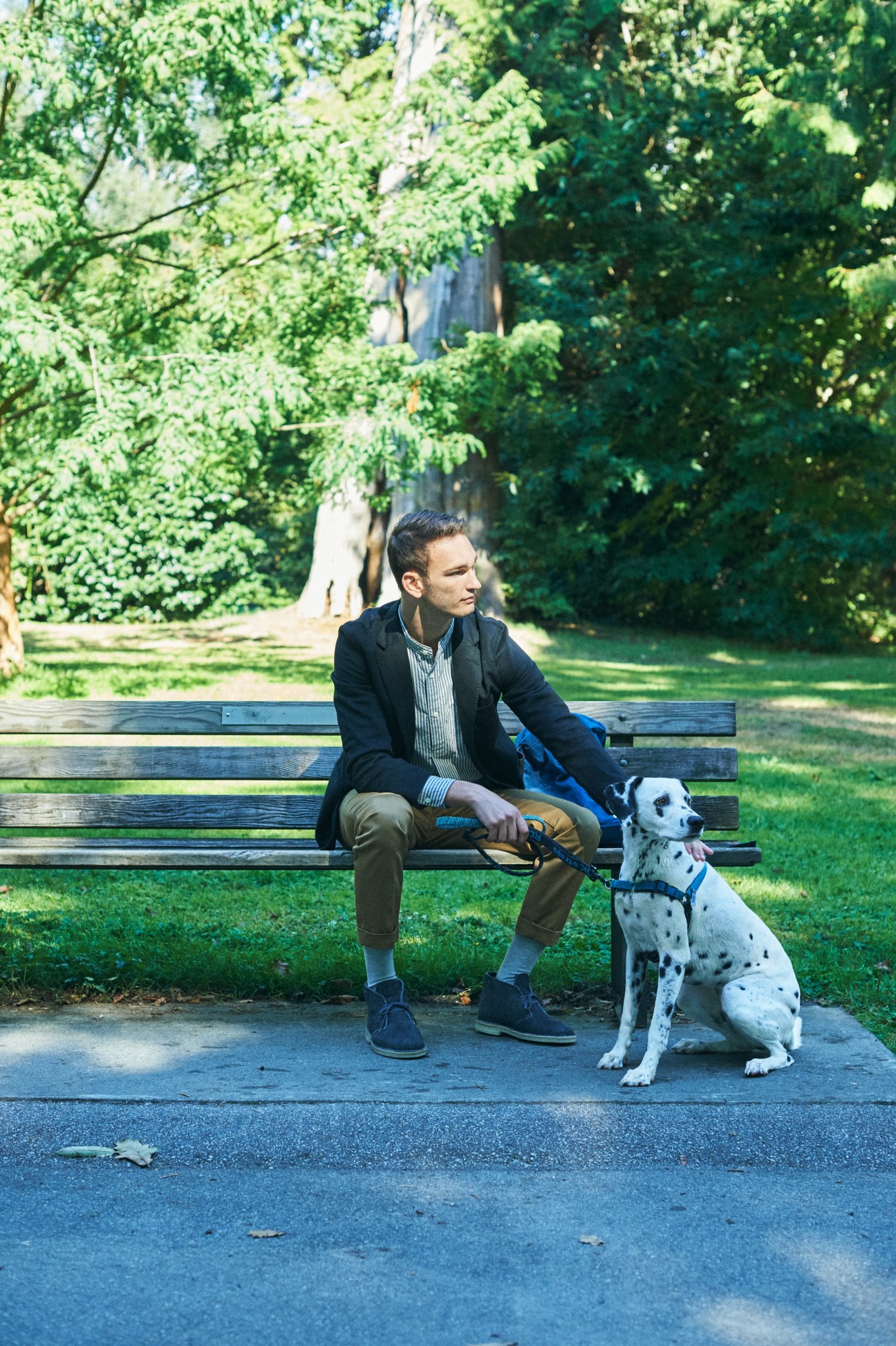 Man in dark blazer and striped shirt sitting on park bench with white spotted dog wearing blue harness