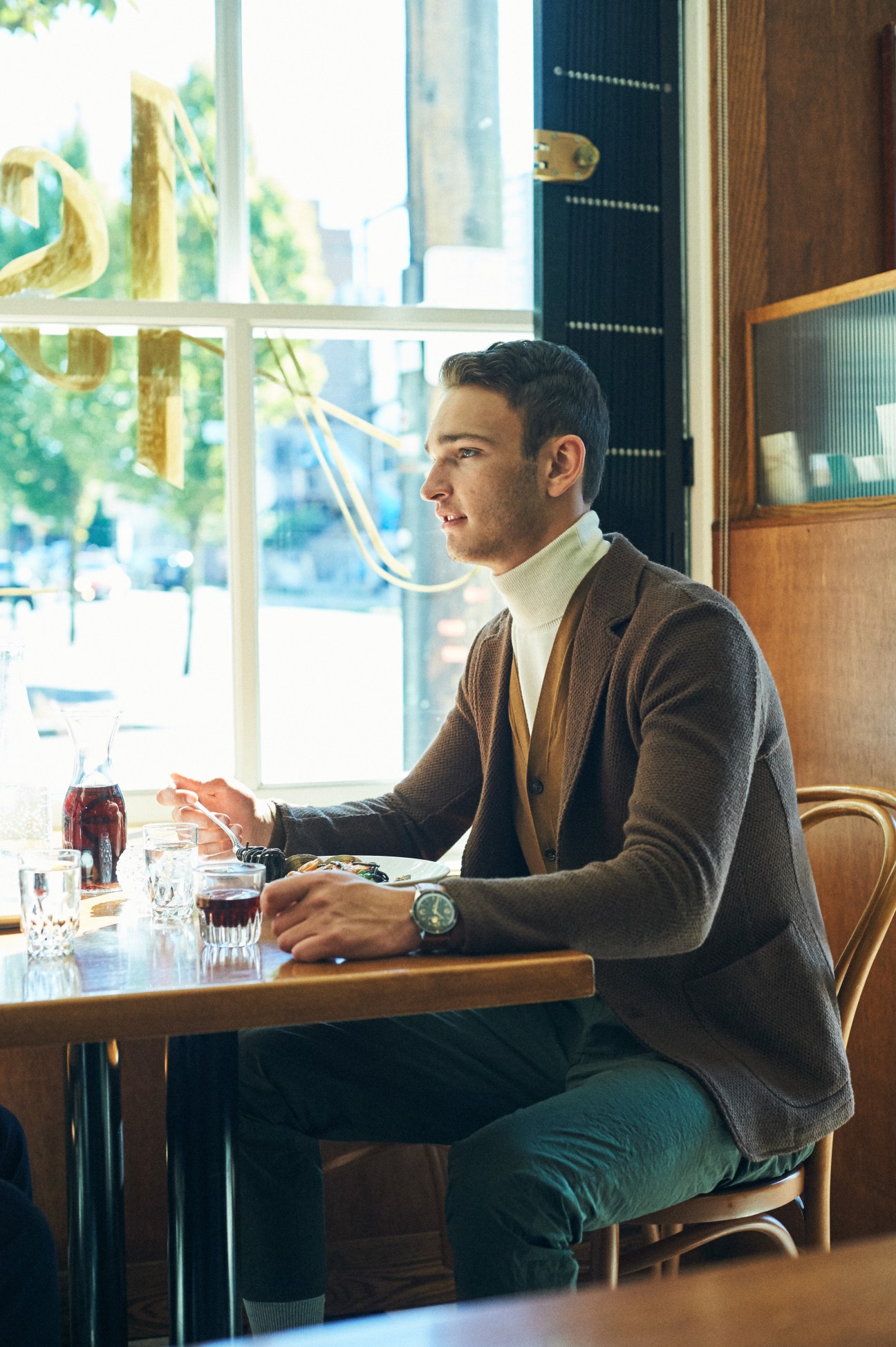 Man in grey cardigan and rollneck jumper sits at cafe table by window with coffee