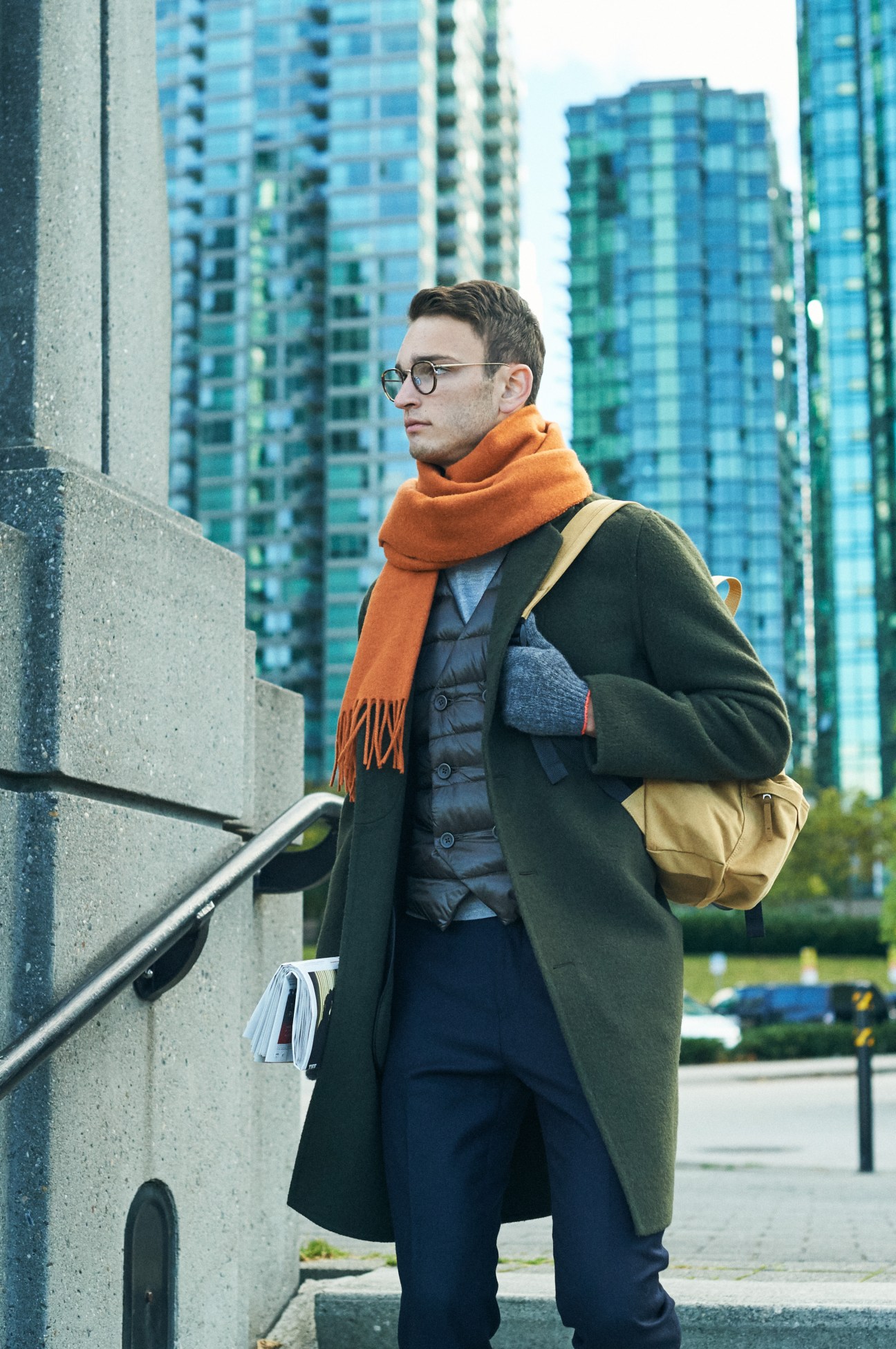 Man in green Belvest coat with orange Wood Wood scarf and yellow bag against Vancouver high-rise buildings