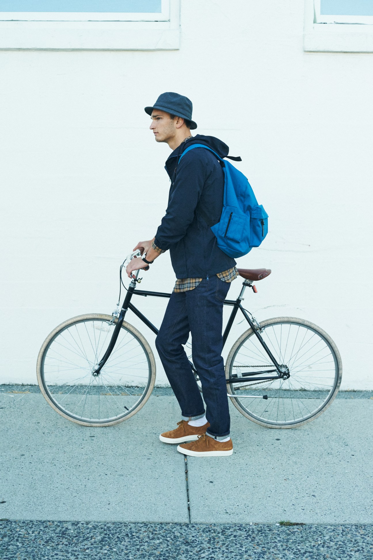 Man with blue backpack and hat standing with black bicycle against white wall