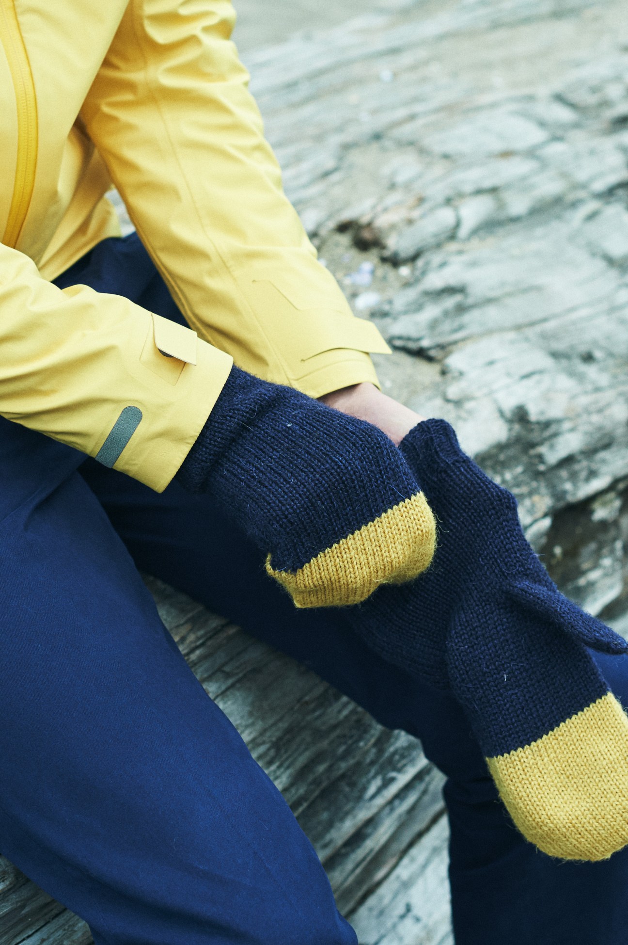 Person wearing yellow jacket and navy blue knit gloves with yellow fingertips seated on wooden dock