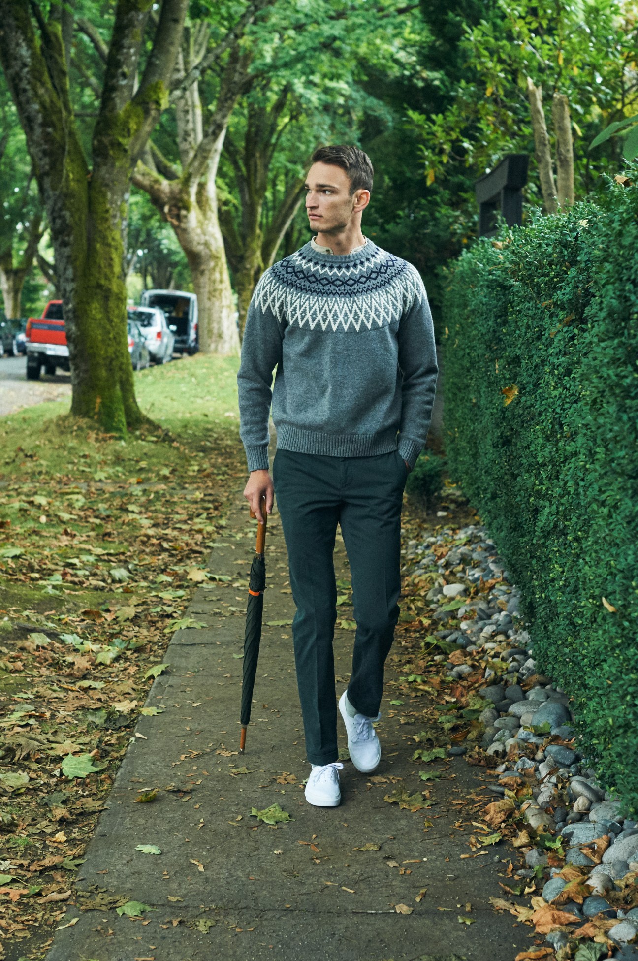 Man wearing grey patterned jumper and dark trousers walking on tree-lined street with umbrella