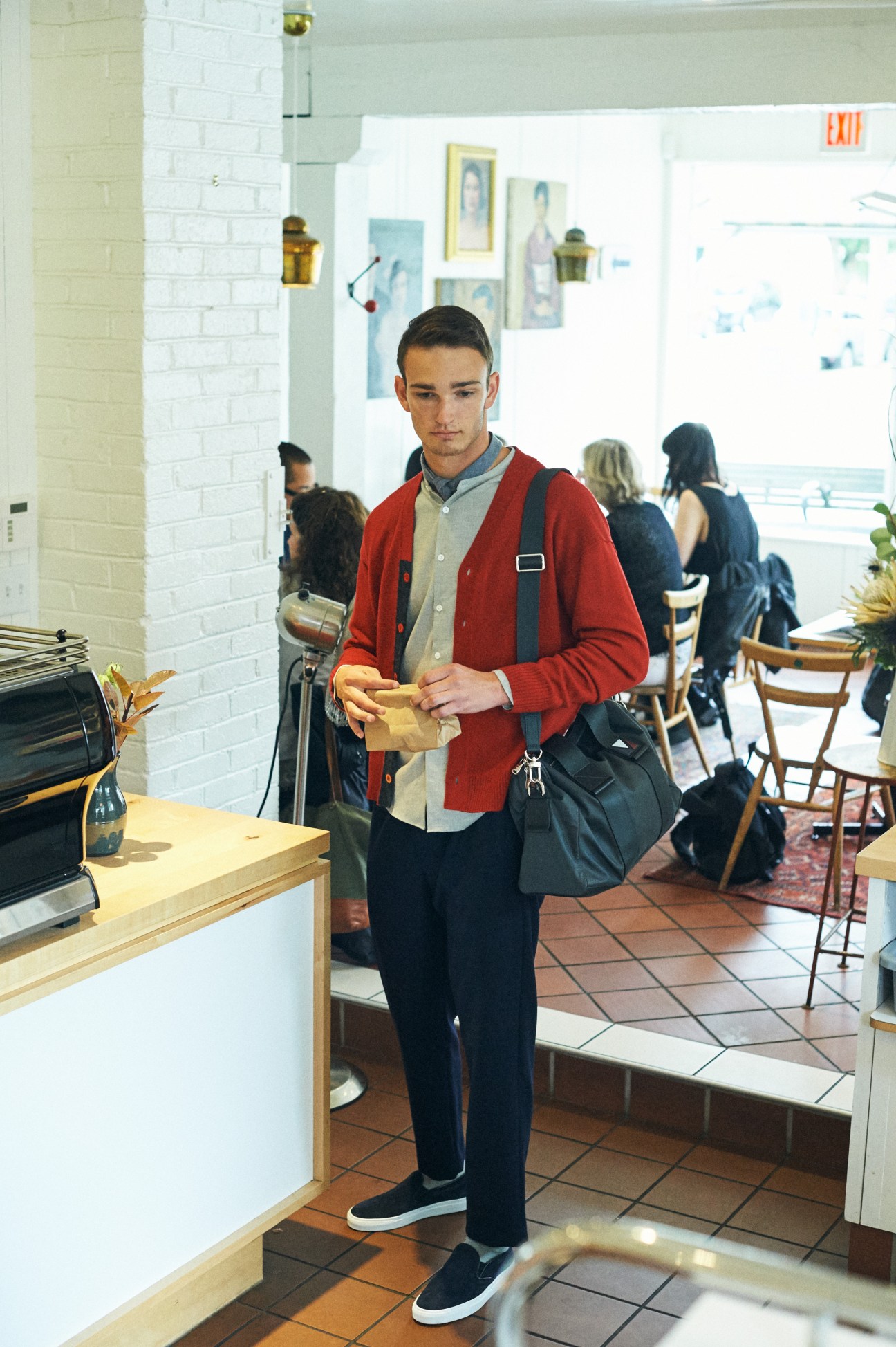 Man in red cardigan holding paper bag in bright cafe with white brick walls and wooden counter