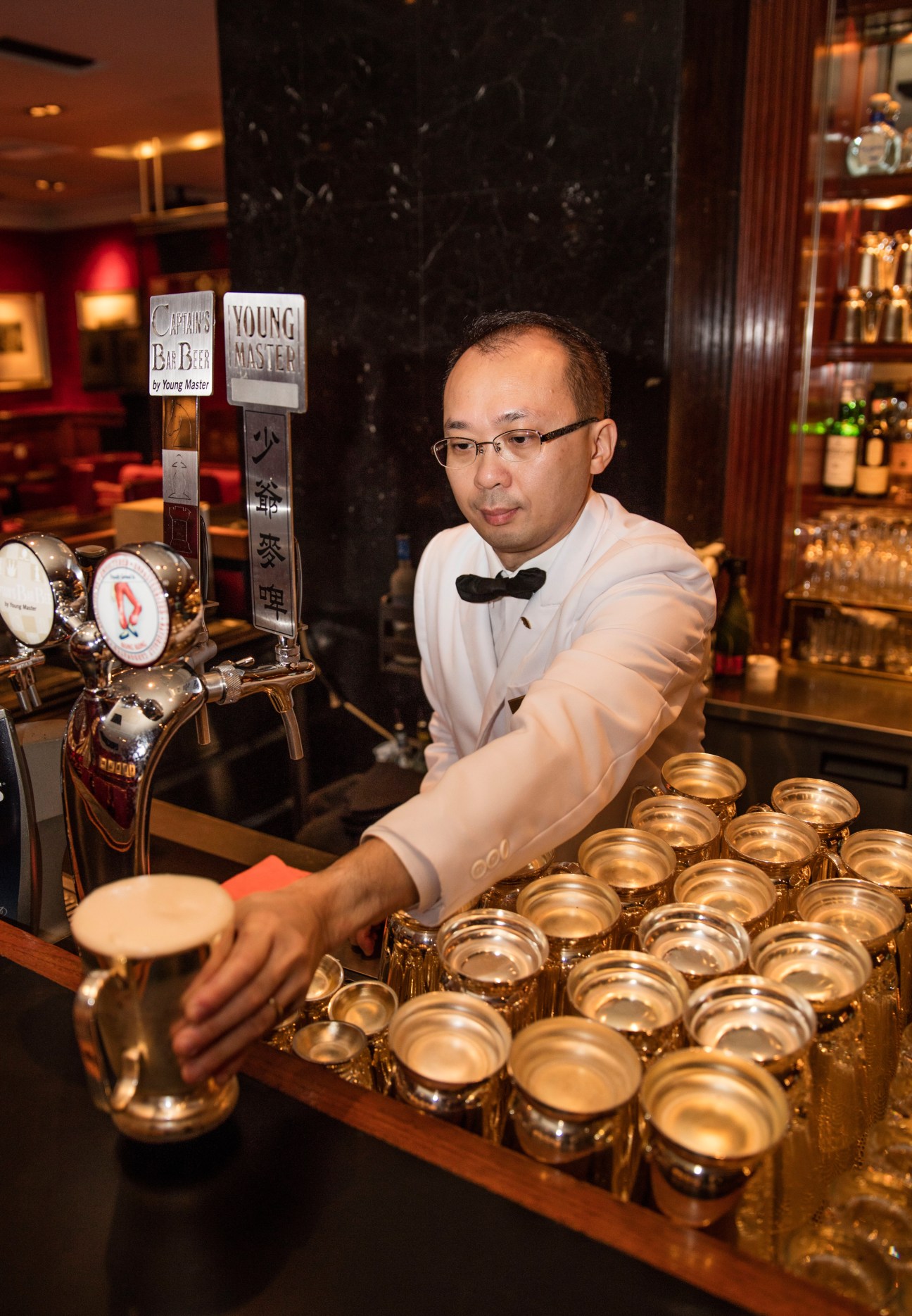 A bartender in white shirt and bow tie pouring drinks behind a bar with multiple glasses lined up.