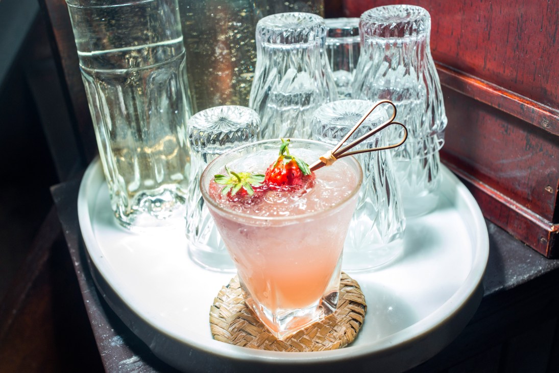 A pink cocktail garnished with fresh strawberry and decorative pick, surrounded by crystal glasses on a white plate at Lola cocktail bar in Tainan.