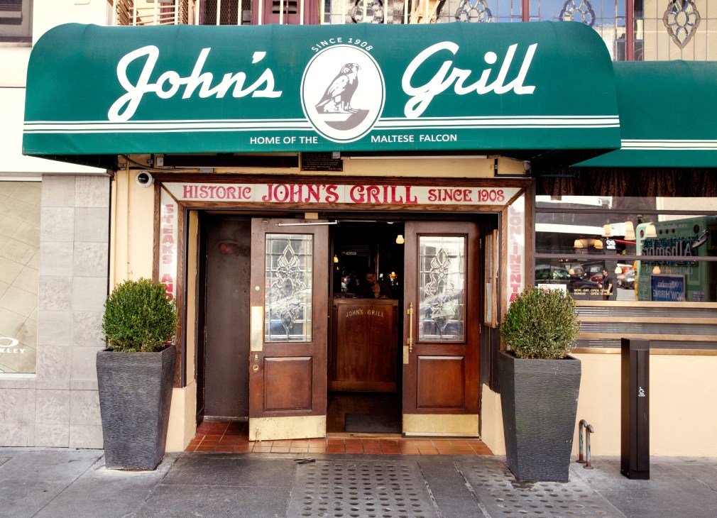 John's Grill storefront with teal awning, established 1908, featuring wood-paneled entrance and potted plants.