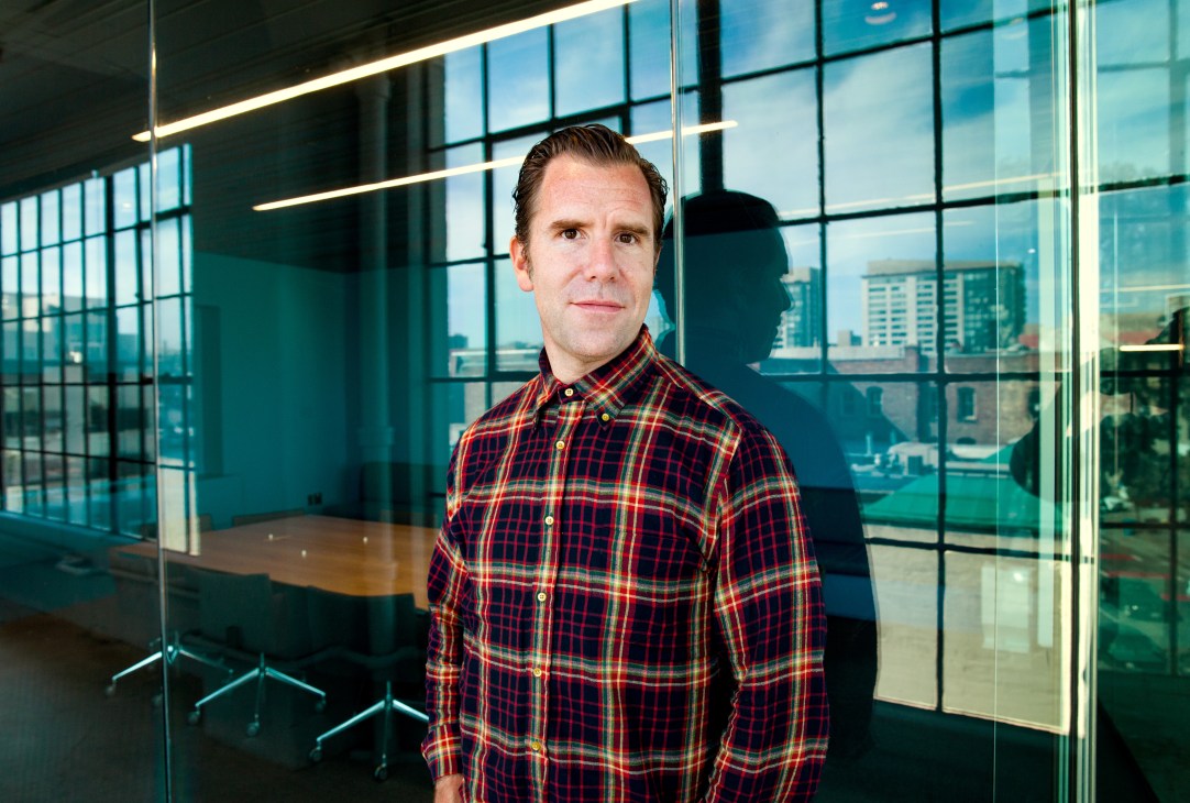Man in red plaid shirt standing in modern office with floor-to-ceiling windows overlooking San Francisco cityscape.