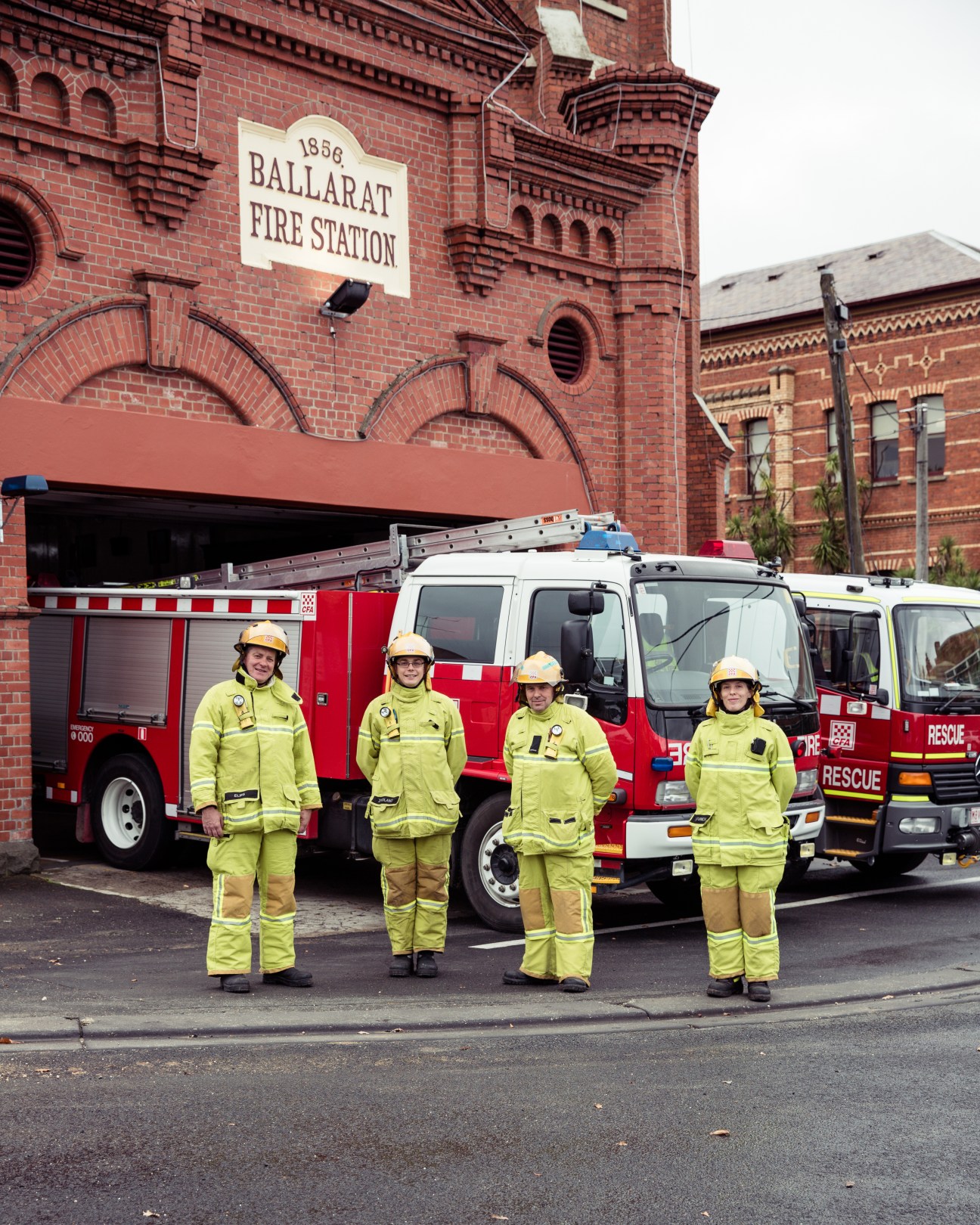 Four firefighters in yellow uniforms stand in front of red fire trucks at the 1856 Ballarat Fire Station in Australia.
