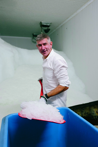 Eric Goeres stands in a freezing packing room at Alaska Ice Corp, holding a bag of ice above a blue bin.