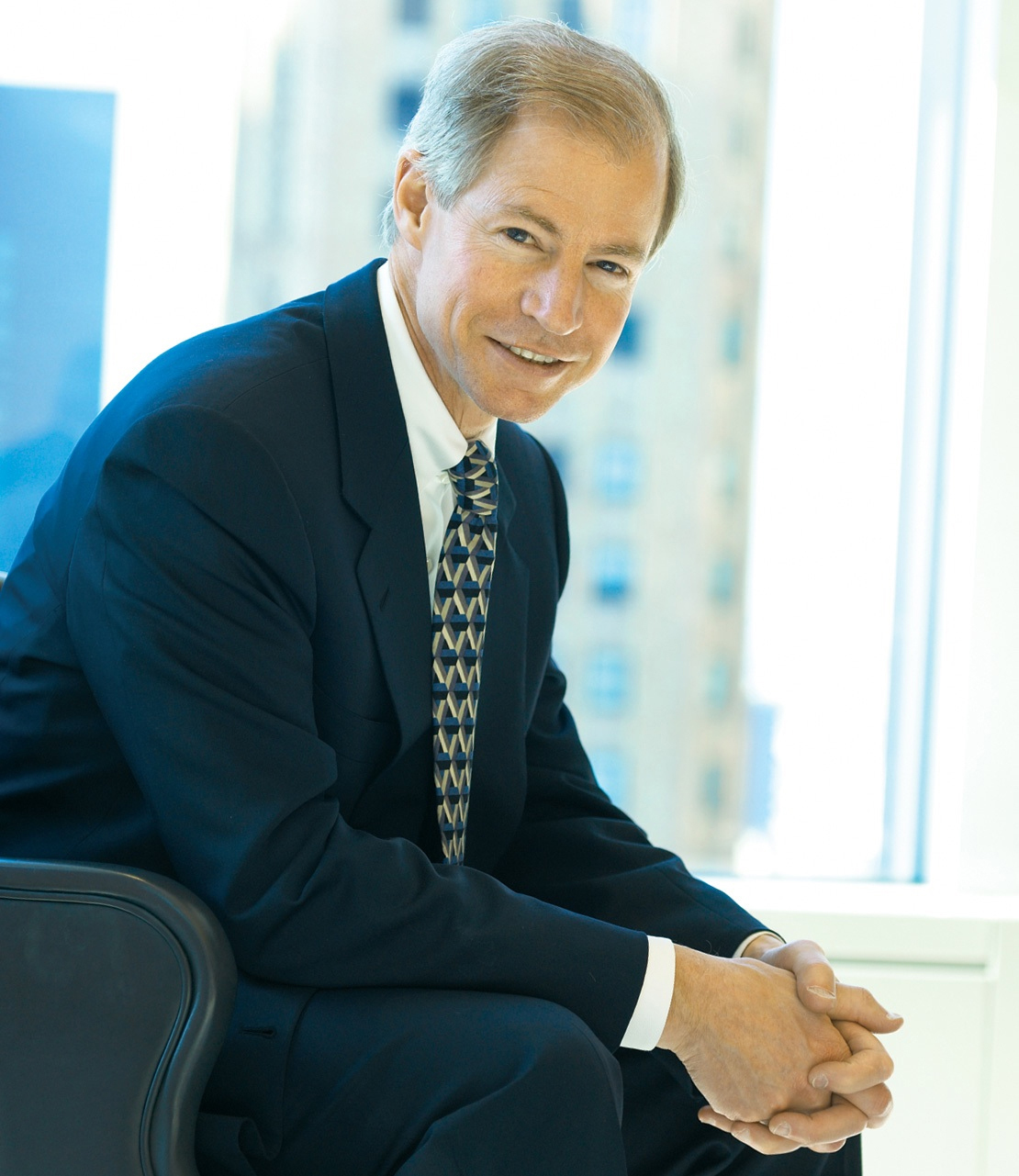 A man in a dark business suit and patterned tie smiles at the camera while sitting indoors.