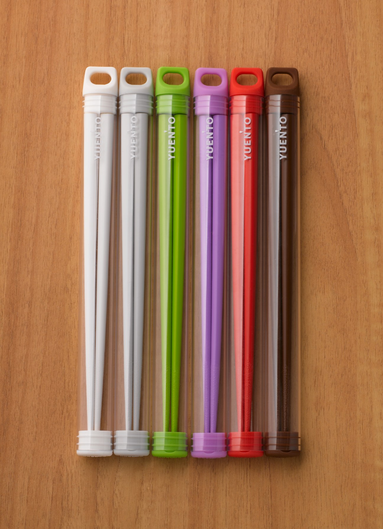 Six Yuen'to Eco Pocke My Hashi portable chopsticks in white, olive, green, purple, red and brown cases arranged in a line on a wooden surface.