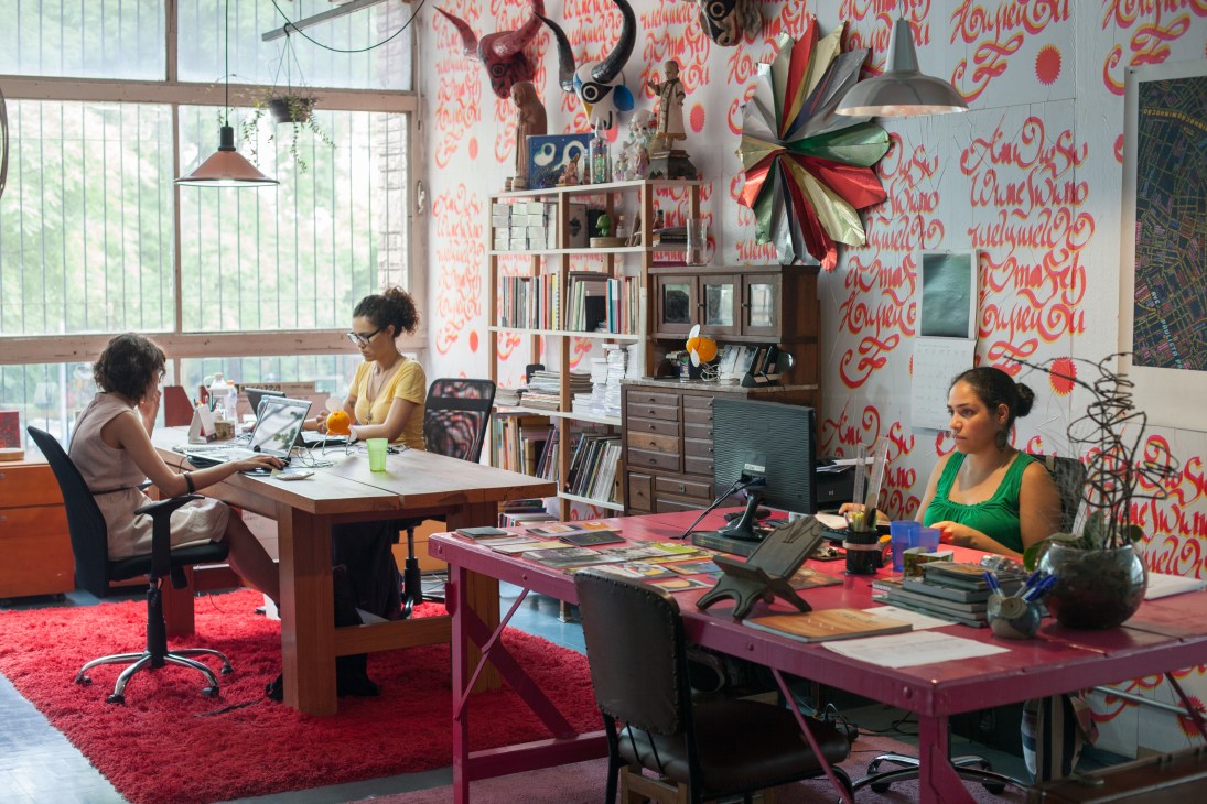 Three people work at desks in a bright office with red patterned wallpaper, shelving, and industrial pendant lighting.
