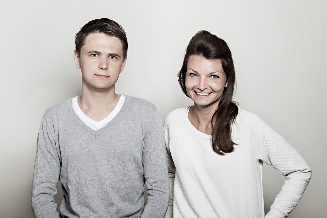Portrait of two young designers, a man in a gray v-neck sweater and a woman in a white long-sleeve shirt, smiling at the camera against a neutral background.