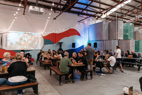 Interior of Colonial Brewing Company in Melbourne with patrons seated at communal tables beneath industrial ceiling and brewery floor visible.