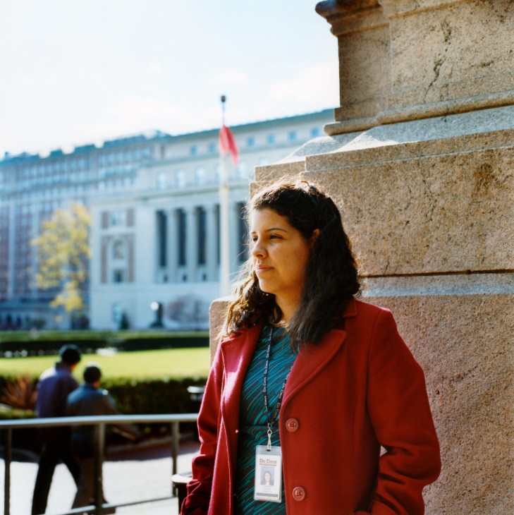 Woman in red blazer with ID badge standing against stone building with classical architecture in background