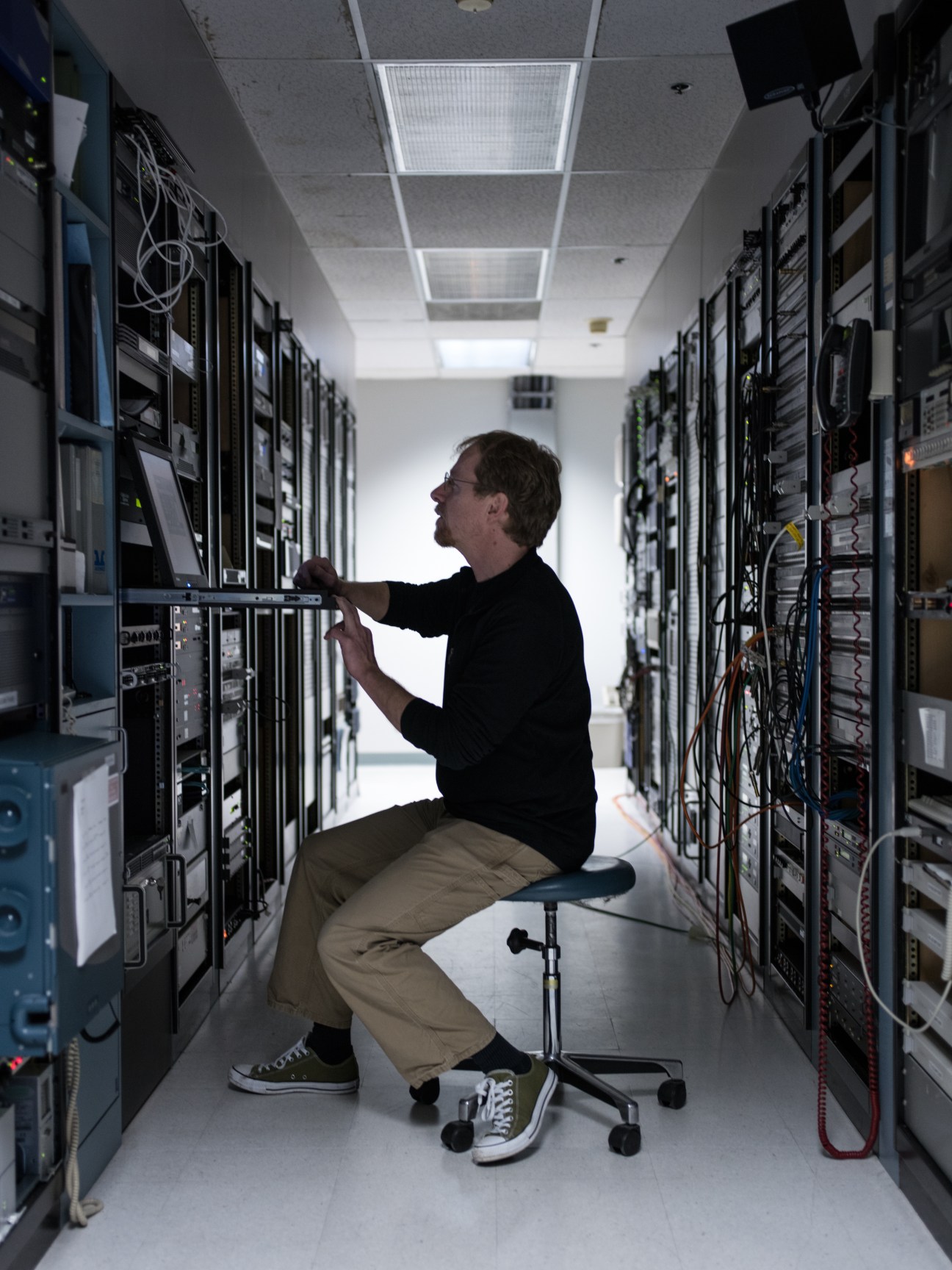 A technician crouches while adjusting equipment in a server room lined with broadcasting infrastructure on both sides.