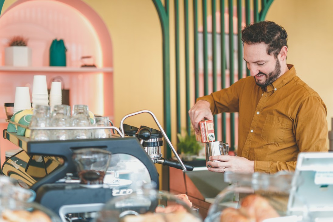 Man in mustard shirt pouring drink at bar counter with pink arched shelving and glassware