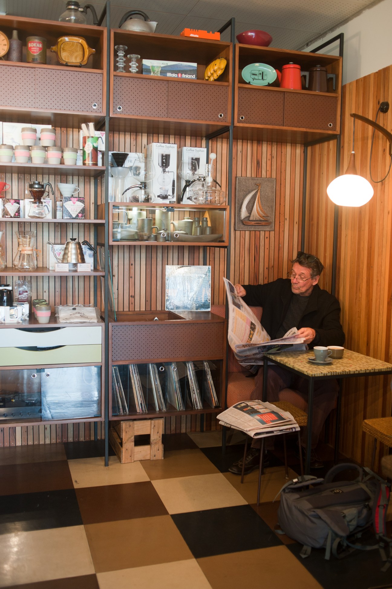 Man reading newspaper at table in mid-century café with wood paneling and shelving.