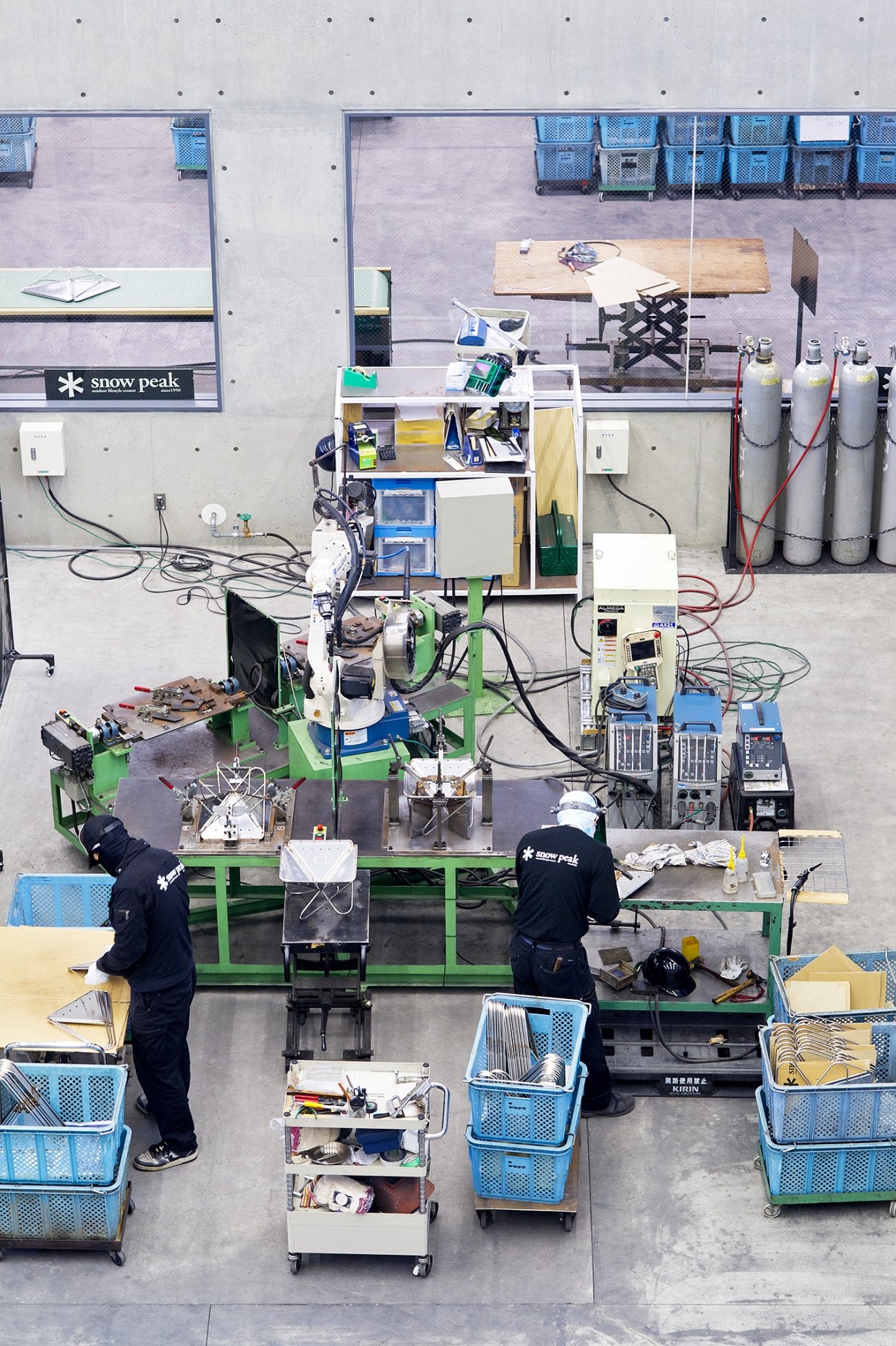 Overhead view of Snow Peak factory floor with two workers assembling products at a green workstation surrounded by blue storage crates and equipment.
