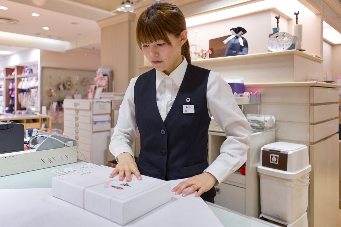 Japanese department store employee gift-wrapping a white box with pink ribbon at Mitsukoshi-Isetan counter.