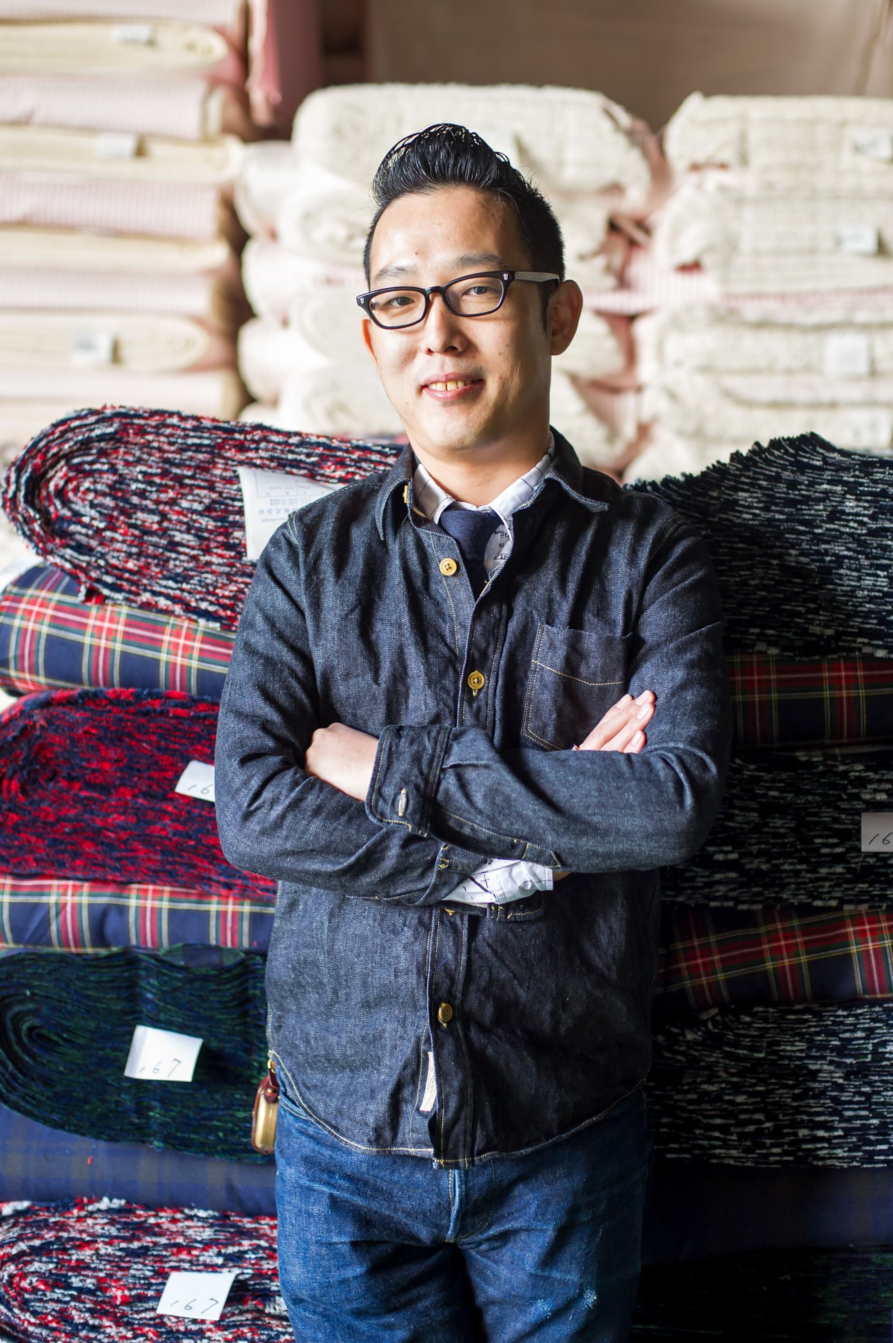 Nobuyuki Ueyama, president of Ueyama Group textile manufacturer Maruwa, standing with arms crossed in front of stacked fabric samples.