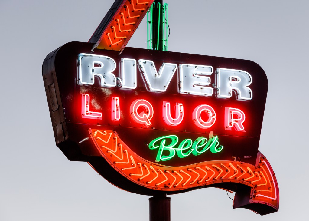 Vintage neon sign reading 'River Liquor Beer' with red and green neon tubes on dark maroon background.