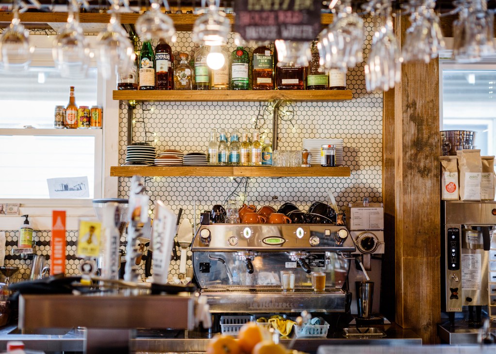 Interior of Quill Kitchen + Cocktails bar with hexagonal tile backsplash, wooden shelves stocked with spirits, and espresso machine.